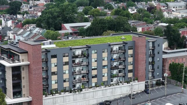 Modern apartment building with green living roof and balconies rises among historic brick rowhouses, parking lot and trees border property in Lancaster, Pennsylvania. Aerial drone view.