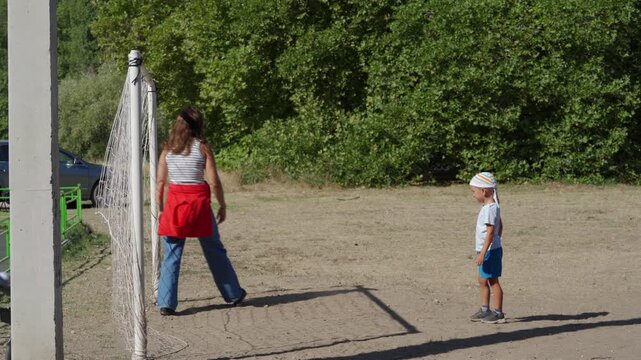 A young boy stands on a dusty field watching his mother retrieve a soccer ball near a net. They enjoy a joyful, carefree day playing outside surrounded by green trees.