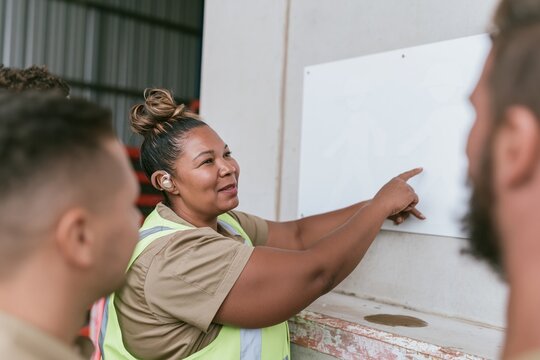 Black woman in hi-vis vest pointing at a safety panel in a cool grey warehouse, natural and vigilant, for EHS training and workplace safety