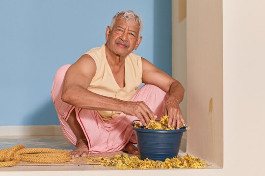 Senior man gathering wilted marigolds into a blue pail on a damp doorstep, showing weathered hands and a dignified expression for waste and reuse