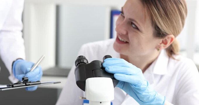 In a bright laboratory, two scientists, focused on their work, peer through a microscope. One woman shares a smile as she points out an intriguing detail, sparking collaboration.