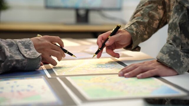 Close-up of military personnel in camouflage uniforms pointing at maps on a table during a tactical planning meeting.