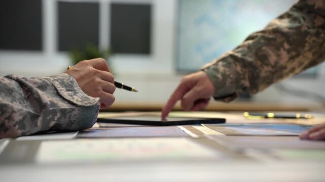 Close view of military personnel in camouflage uniforms discussing documents and pointing at maps on a desk during a planning meeting.