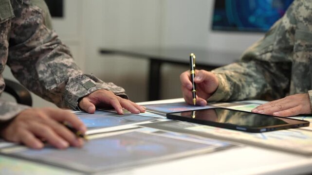 Close-up of military personnel in camouflage uniforms reviewing maps and digital tablets during a planning meeting at a table.