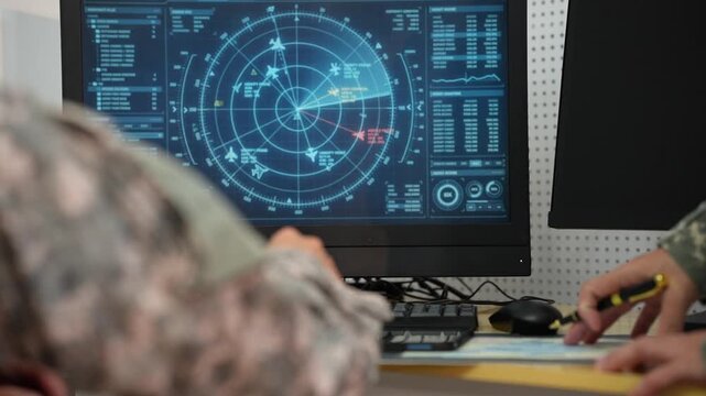 Military personnel in camouflage monitor a radar tracking display at a control desk, showing aircraft positions, data charts and digital surveillance information.