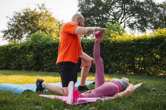 Athletic man assisting woman with hamstring stretch on fitness mat during couple workout in sunny park, outdoor training concept