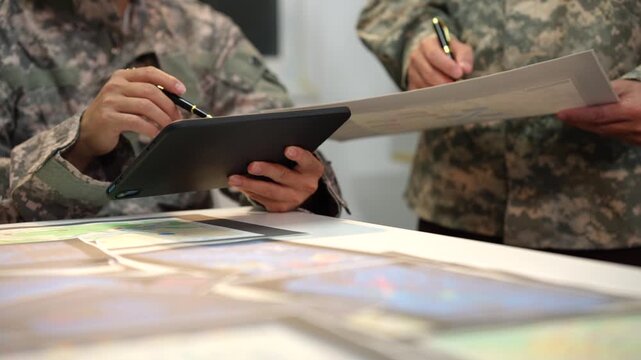 Close-up of military personnel in camouflage uniforms reviewing papers and using a tablet during a planning meeting at a table.