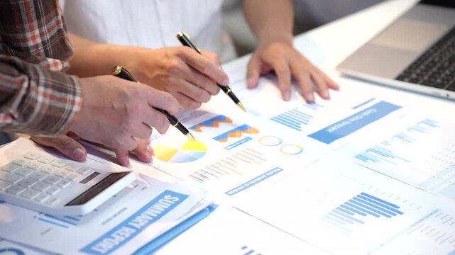 Close-up of business colleagues reviewing colorful financial charts and reports on a desk with calculator, pen, and laptop during a planning meeting.