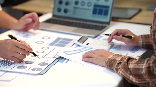 Close-up of coworkers reviewing printed business charts, reports and calculator data beside a laptop during an office meeting.