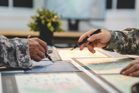 Close-up of military personnel in camouflage uniforms pointing at maps and documents during a planning meeting at a desk.