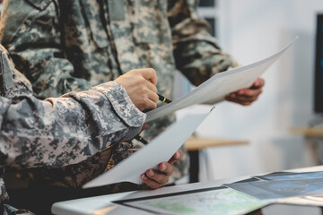 Close-up of military personnel in camouflage uniforms reviewing documents and maps at a table during a planning meeting. © NanSan