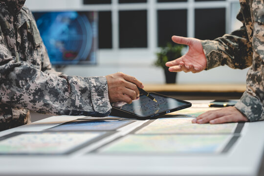 Two military personnel in camouflage uniforms use a tablet over maps during a planning meeting in a modern office.