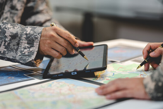 Close-up of military personnel using a tablet and paper maps with pens during a planning meeting, showing teamwork, navigation, and field strategy.