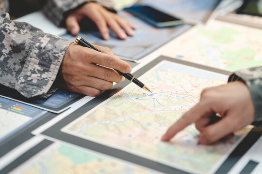 Close-up of military personnel planning a route on printed maps with a tablet and digital documents on a table.