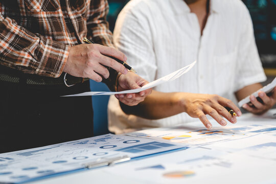 Business colleagues review printed charts and reports at a desk, pointing at financial data during a meeting and planning session.