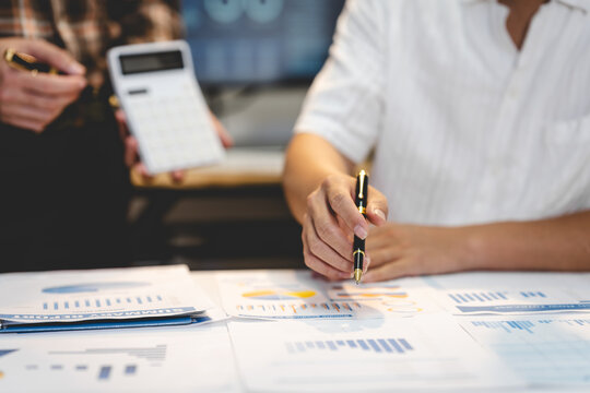 Business professionals review financial charts and paperwork at a desk, with one person writing notes and another holding a calculator.