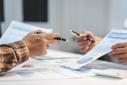 Close-up of two business people discussing financial charts and reports at a desk, pointing with pens beside a calculator in a bright office.