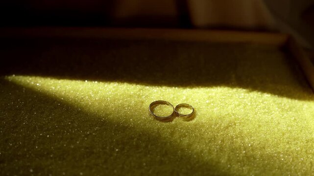 Two gold wedding rings resting on glittery yellow granular surface inside shallow tray under narrow warm beam of light, with ring, gold, wedding, jewelry detail