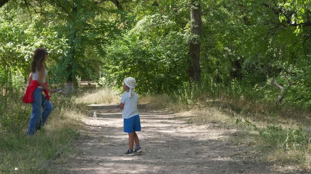 A mother walks along a dirt path as her young son stands watching her. They enjoy playing badminton together in the shady forest. They feel happy, peaceful, and relaxed.