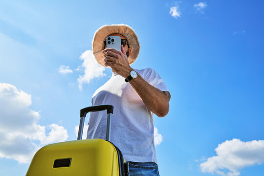 Low Angle View of Male Traveler with Yellow Suitcase Taking Photo with Smartphone Under Blue Sky