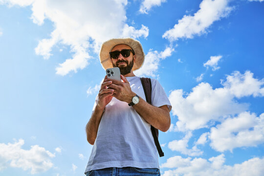 Male tourist in hat and sunglasses using smartphone outdoors against blue sky
