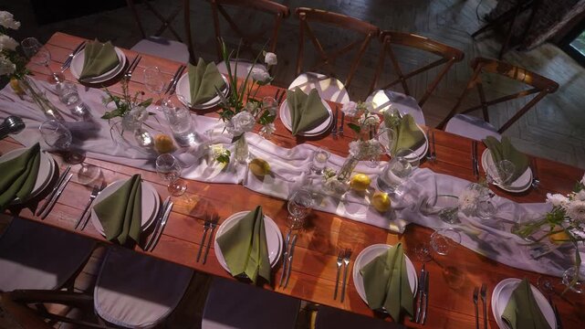 Top-down banquet table set with folded olive green napkins on white plates and glass centerpieces, with table, dining, wood, plate detail, showing napkin, cutlery, glass, vase context