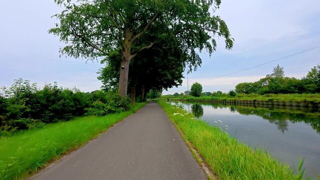 First-person view moving forward on a paved path next to a tranquil canal, surrounded by lush green trees and nature.