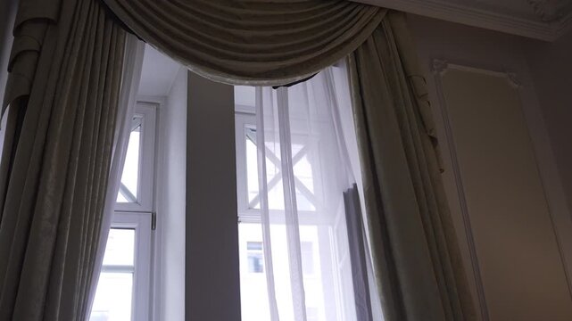 Ornate upper window with layered drapery and subtle sheer movement under diffused daylight, tall interior windows framed by pleated side curtains and semicircular swag valance