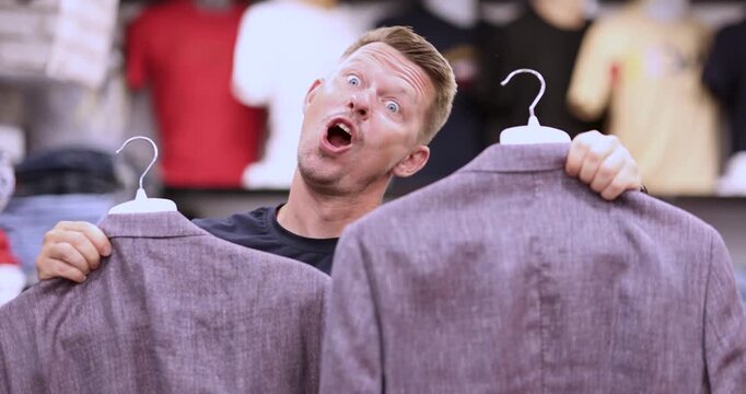 In a lively thrift shop filled with colorful clothes, a man joyfully showcases two stylish jackets. His excitement is contagious as he grins widely, celebrating his find in this treasure trove.