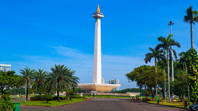 The iconic National Monument (Monas) in Jakarta, Indonesia, is surrounded by a spacious plaza, green landscapes, palm trees, and modern city buildings beneath a bright blue sky. 