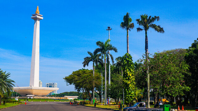 The iconic National Monument (Monas) in Jakarta, Indonesia, is surrounded by a spacious plaza, green landscapes, palm trees, and modern city buildings beneath a bright blue sky. 