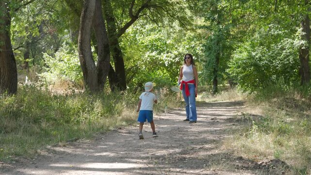 A caring mother and her happy son stand holding badminton rackets. They share a joyful, peaceful game on a dirt path in a green park.