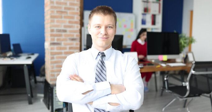 A man in a white shirt and tie poses confidently with arms crossed, surrounded by a modern office. A colleague is visible working in the background, creating a lively atmosphere.