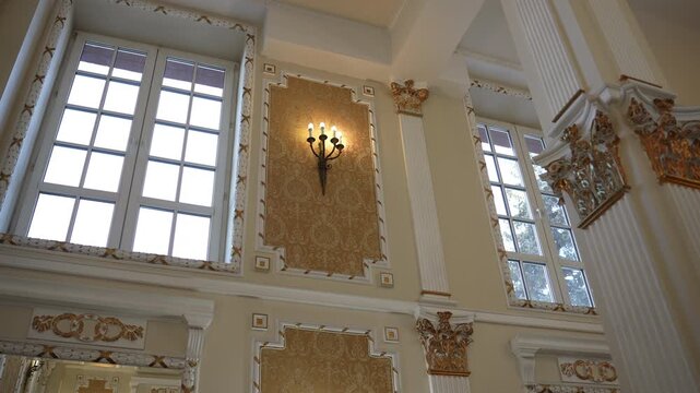 Low-angle view of ornate classical hall interior with illuminated wall sconce, tall mullioned windows, gilded mouldings, carved details, warm glow, elegant symmetry
