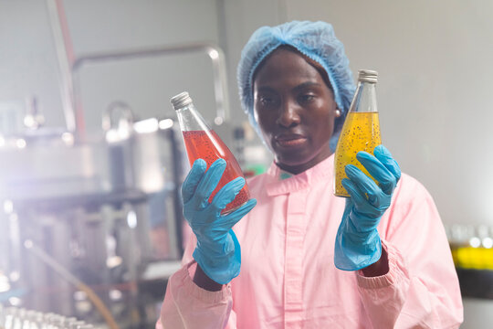 Black woman inspecting drink quality with rows of glass bottles on the automated conveyor system in manufacturing assembly line.