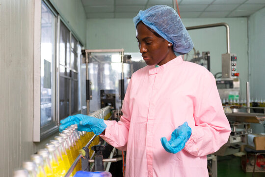 Black woman inspecting drink quality with rows of glass bottles on the automated conveyor system in manufacturing assembly line.