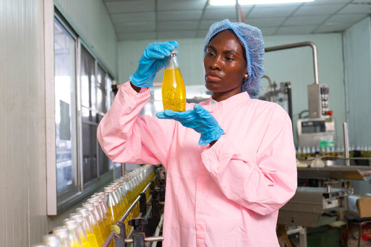 Black woman inspecting drink quality with rows of glass bottles on the automated conveyor system in manufacturing assembly line.