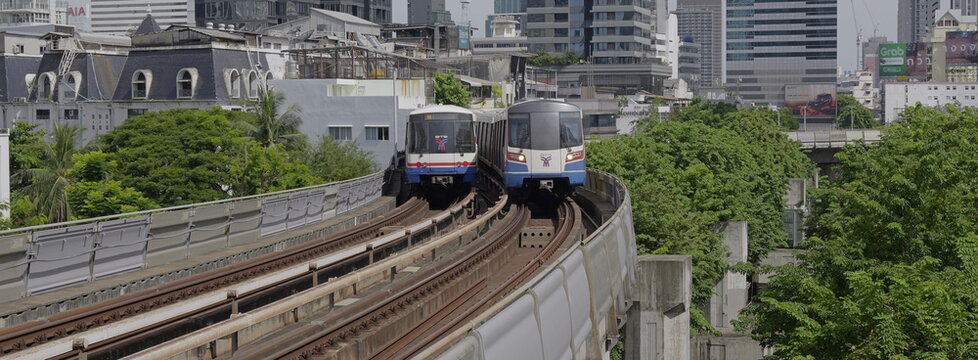 Two elevated BTS Skytrain passenger trains passing each other on curved railway tracks against an urban skyline background in Thai capital Bangkok