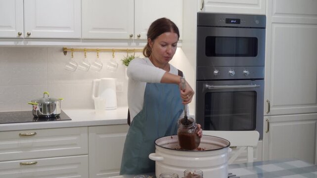 A focused woman scoops rich homemade sauce into glass jars from a big pot. She works with care in a bright, cozy kitchen. The calm atmosphere brings a feeling of warm satisfaction.