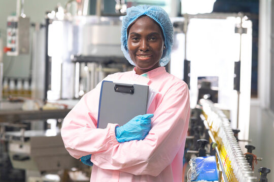 Black factory nutritious woman inspecting drink quality with rows of glass bottles filled with nutritious health drinks on the automated conveyor system in manufacturing assembly line.
