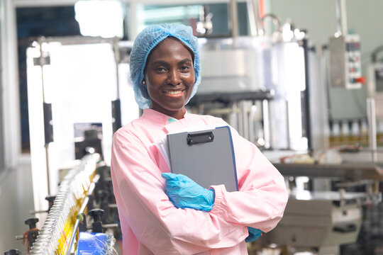 Black factory nutritious woman inspecting drink quality with rows of glass bottles filled with nutritious health drinks on the automated conveyor system in manufacturing assembly line.