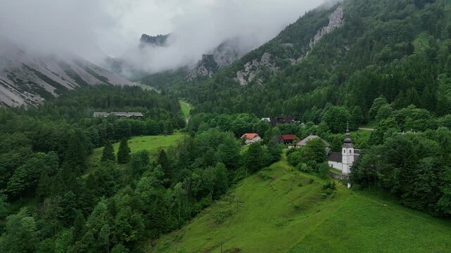 Drone passes by Catholic church in Slovenia Alps near Austrian border, during cloudy summer day with dramatic mountain backdrop and green forest creating cinematic travel perspective