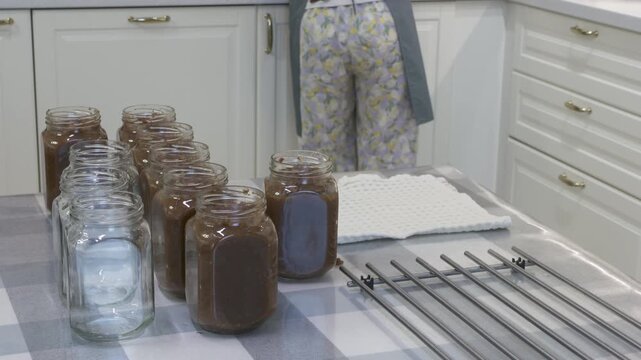 Rows of glass jars sit filled on a kitchen table while a woman works quietly nearby. The clean white room feels peaceful and cozy. Her completed work brings a sense of warm satisfaction.