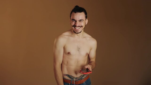 Shirtless young man with topknot hairstyle holds and interacts with red smartphone while shifting from questioning gaze to broad smile and finally looking down at device, expression progression
