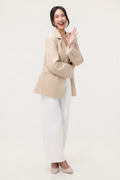 Happy young Asian businesswoman showing OK hand sign gesture with a bright smile, corporate studio portrait on white background.