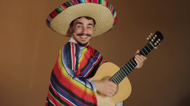 Young man wearing wide-brimmed straw sombrero with colorful woven trim and striped serape plays acoustic guitar in front of plain warm brown studio backdrop, with man, guitar, sombrero, serape detail