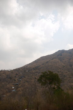 Vertical view from the ground looking up at the steep mountain slope and pilgrimage trail with local shops among dry forest trees under an overcast cloudy sky at Khao Wong Phrachan in Lopburi Thailand