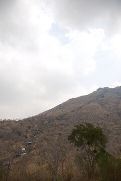 Vertical view from the ground looking up at the steep mountain slope and pilgrimage trail with local shops among dry forest trees under an overcast cloudy sky at Khao Wong Phrachan in Lopburi Thailand
