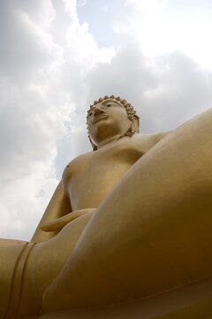 Dramatic low angle view looking up past the massive golden seated legs towards the serene face of the giant Buddha statue under bright white clouds in the sky at Khao Wong Phrachan in Lopburi Thailand