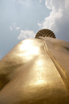 Rear low angle view looking up at the sunlit golden back shoulder and head of the giant Buddha statue shining brightly under a blue sky with soft white clouds at Khao Wong Phrachan in Lopburi Thailand